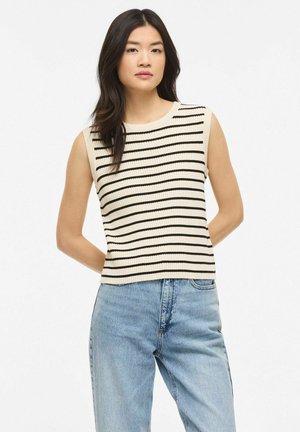 Jeune femme aux longs cheveux foncés porte un pull sans manches beige avec des rayures horizontales noires et un jean taille haute bleu clair, debout les mains derrière le dos.