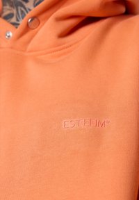Haut à capuche orange en tissu doux, avec une poche frontale, des boutons-pression et un logo « ESTEEM » en relief de la même couleur.