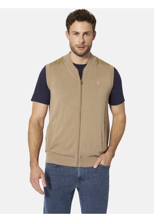 Mann trägt beige ärmellose Weste mit Reißverschluss über marineblauem Kurzarmhemd und blauen Jeans, steht mit einer Hand in der Tasche vor weißem Hintergrund.