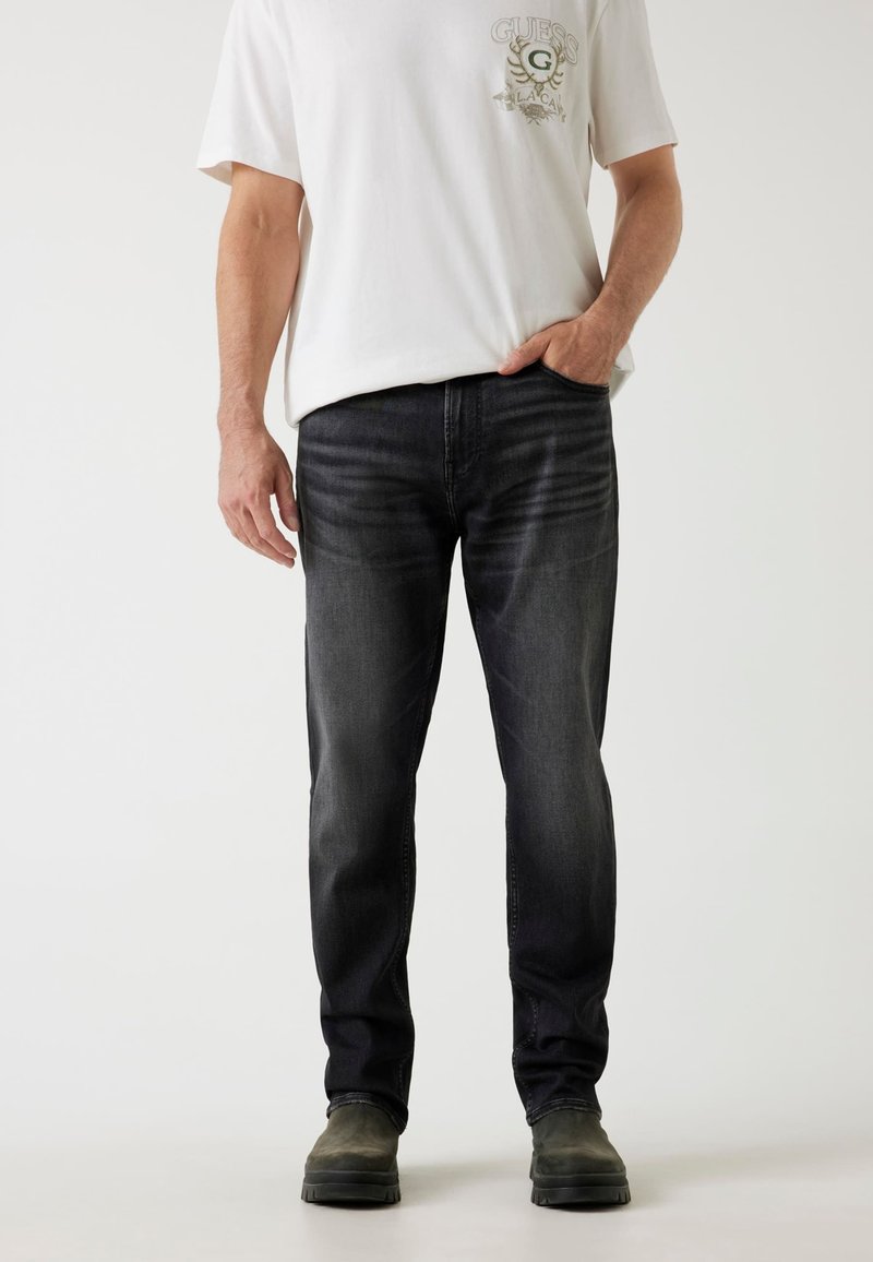 Dunkelgraue Jeans mit geradem Beinschnitt, mittlerer Taillenhöhe und subtilen Verwaschungen, kombiniert mit einem weißen Grafik-T-Shirt und dunklen Schuhen.