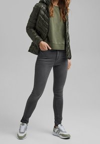 Puffig mörkgrön jacka med huva, olivgrön sweatshirt, grå tajta jeans och vita sneakers med gröna accenter. Släta, quiltade texturer.