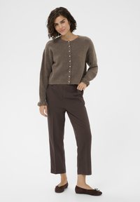Brown, textured cardigan with front buttons, worn over brown pants; model poses in flat brown shoes. Simple design with a soft finish.