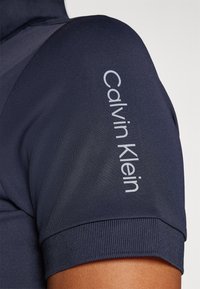 Tumesinise polo särk ribatud varrukatega, millel on silmatorkav valge "Calvin Klein" logo varrukal. Valmistatud tekstuurilisest, elastsemast kangast.