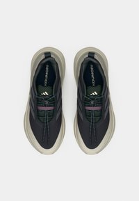 Μαύρα και γκρι αθλητικά παπούτσια με υφασμένη επάνω επιφάνεια, πράσινες και μωβ λεπτομέρειες, και χοντρή μπεζ σόλα. Διαθέτουν ελαστικά κορδόνια και λογότυπο.