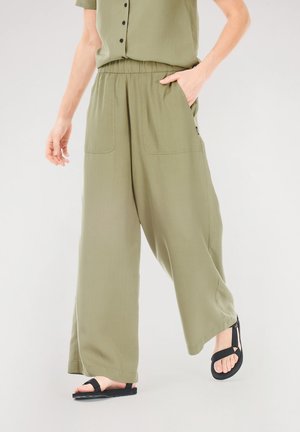 Weite Hosen aus leichtem, olivgrünem Stoff. Ausgestattet mit einem elastischen Bund, Seitentaschen und einem fließenden Design. Kombiniert mit schwarzen Sandalen.