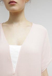 Haut rose clair à col V avec des manches courtes, fabriqué à partir d'un tissu texturé. Une couche intérieure blanche est partiellement visible sous le haut.