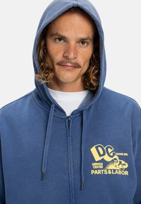 Námornícky modrá mikina na zips s kapucňou na šnúrky. Obsahuje žltý grafický logotyp s nápisom "DG Shoe Co. Servisné stredisko Diely & Práca."