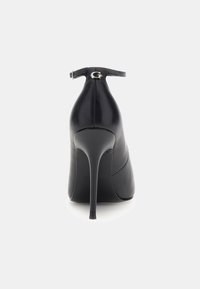 Chaussure noire en cuir à talon haut avec un talon aiguille mince et une lanière de cheville, présentant un détail de fermoir en métal à l'arrière.