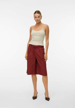 Femme debout portant un top en maille beige sans manches, un short bordeaux taille haute à jambes larges et des talons pointus foncés sur fond blanc.