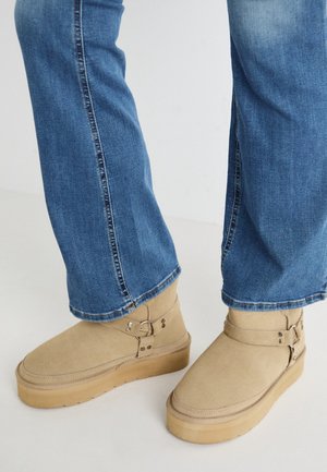 Personne portant un jean bleu et des bottes en daim beige avec des brides à boucle, debout sur une surface simple.