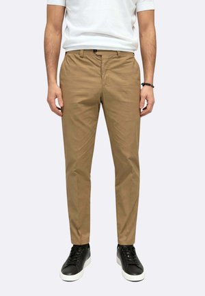 Uomo che indossa pantaloni color cuoio aderenti, camicia bianca a maniche corte, braccialetto nero e scarpe da ginnastica nere con suola bianca, in piedi davanti a uno sfondo semplice.