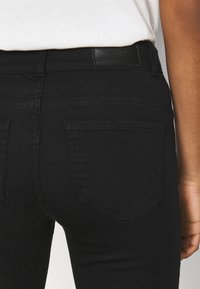 Jeans en denim noir avec une texture lisse, présentant des poches arrière et une étiquette rectangulaire à la taille. Vue rapprochée sur le côté.