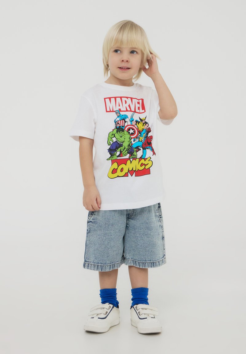 Maglietta bianca con personaggi colorati dei Marvel Comics, abbinata a calzini blu e pantaloncini di jeans. Un bambino sta in piedi su uno sfondo neutro.