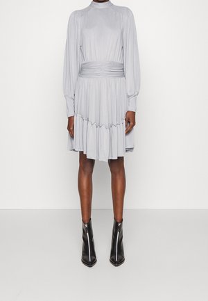 Femme portant une robe plissée à manches longues gris clair avec un col montant et une jupe à volants, associée à des bottines noires à bout pointu.