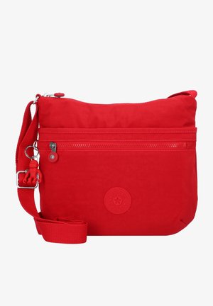 Red fabric crossbody bag with zippered pocket, adjustable strap, and a round logo patch. Smooth texture with a small monkey keychain attached.