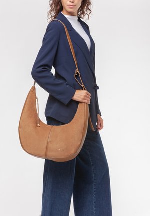 Donna in blazer blu navy e jeans a gamba larga che tiene una grande borsa a tracolla in suede beige con tracolla regolabile su sfondo bianco.