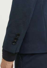 Manica di un blazer blu navy con un tessuto testurizzato, tre bottoni neri e un dettaglio a costine per un contrasto aggiunto.