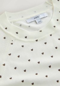 Pull blanc avec un col côtelé, présentant un motif de petits cœurs bruns espacés uniformément sur le tissu. Étiquette de la marque visible.