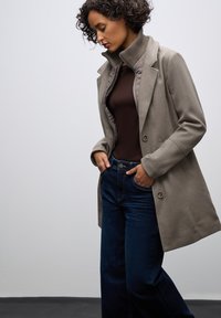 Cappotto in misto lana beige con un ampio colletto, bottoni doppi, e foderato trapuntato, abbinato a jeans in denim scuri e a un top marrone.