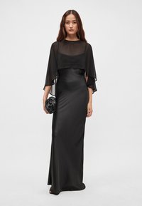 Černé večerní šaty s průsvitnou vrstvou, přiléhavým střihem, dlouhými rukávy, hladkou texturou a sbíranou kabelkou. Minimalistický design.