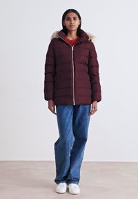 Bordo puf jakna s kapuljačom i umetkom od umjetnog krzna, koja ima prednji zatvarač, quilted teksturu i dugačke rukave, u kombinaciji s raskošnim plavim trapericama.