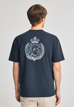 Donker marineblauwe katoenen t-shirt met een groot wit logo op de achterkant, verfraaid met een kroon en lauriertakken. Korte mouwen, relaxte pasvorm.