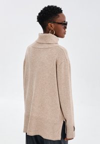 Beżowy sweter z golfem o luźnym kroju, z prążkowanymi mankietami i bocznymi rozcięciami. Miękka tekstura, z zagiętym wzorem kołnierza.