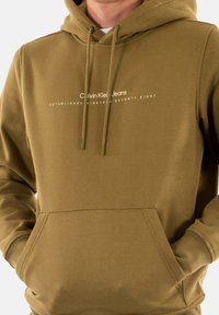 Sudadera de algodón verde olivo con un bolsillo frontal, capucha con cordón y logo impreso en blanco que dice "Calvin Klein Jeans", centrado sobre el bolsillo.