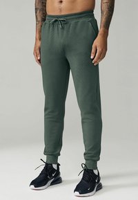 Green, tapered joggers made of textured fabric with an elastic waistband and drawstring, featuring side pockets and ribbed cuffs.