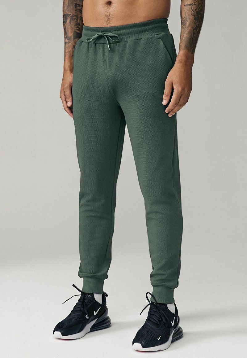 Green, tapered joggers made of textured fabric with an elastic waistband and drawstring, featuring side pockets and ribbed cuffs.