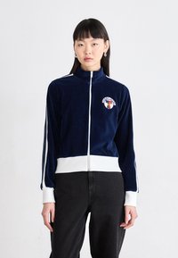 Giacca zip-up in velluto blu navy con dettagli bianchi e logo, caratterizzata da un colletto alto e strisce laterali, abbinata a pantaloni neri.
