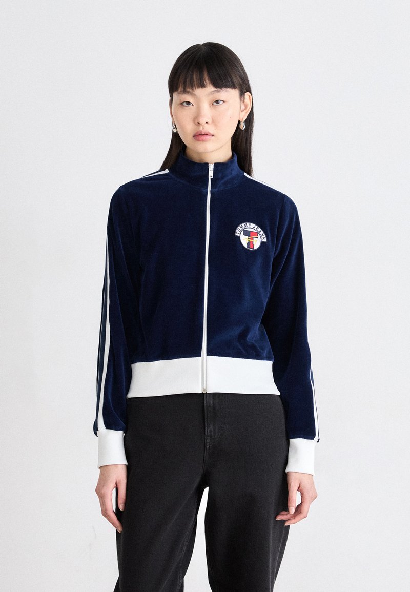 Giacca zip-up in velluto blu navy con dettagli bianchi e logo, caratterizzata da un colletto alto e strisce laterali, abbinata a pantaloni neri.