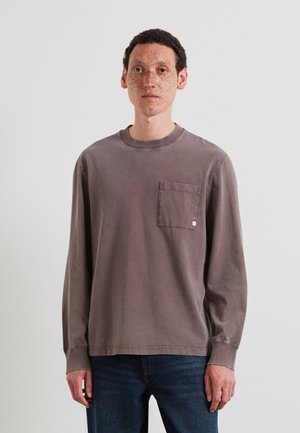 Uomo con capelli corti e ricci e lentiggini, che indossa una maglia grigia a maniche lunghe con taschino sul petto e jeans blu, in piedi davanti a uno sfondo semplice.