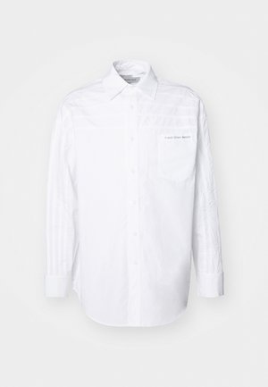 Biała koszula z długim rękawem wykonana z bawełny. Posiada zapięcie na guziki z przodu, kołnierz oraz kieszeń na lewej piersi z subtelnym logo. Teksturowane panele boczne.