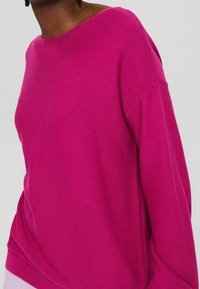 Personne portant un pull à manches longues rose vif avec un large col rond sur un fond clair uni.