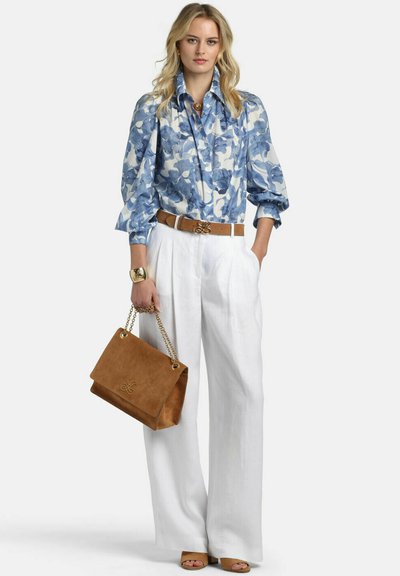Woman wearing blue floral blouse, white wide-leg pants, tan belt, tan handbag with gold chain, gold bracelet, and tan open-toe heels, standing.