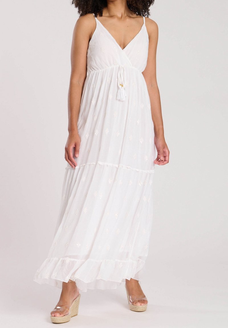 Femme portant une robe maxi blanche sans manches avec des motifs brodés et un lien à la taille, associée à des sandales compensées transparentes.