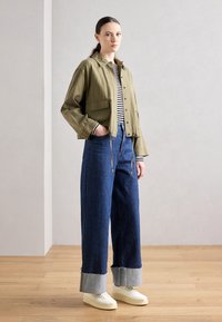 Veste courte olive avec boutons-pression, haut rayé, jean bleu à pattes larges avec ourlet retroussé, et baskets blanches.