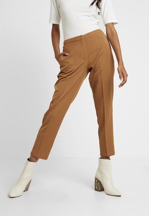 Woman wearing brown tailored ankle-length pants, white top, and white ankle boots with snakeskin-patterned heels, posing with one hand in pocket.