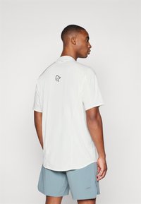 Hvit sportsshirt med korte ermer og rund hals, med en liten svart logo på ryggen. Glatt tekstur, lett design.