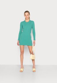 Vestido azul-turquesa de mangas longas com um corte ajustado, combinado com sapatos de salto amarelos e uma pequena bolsa amarela. Fundo simples.