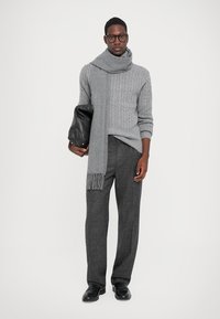 Mand iført en grå kabelstrikket sweater, matchende tørklæde, mørkegrå bukser, sorte sko og briller, mens han holder en sort lædertaske.