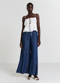 Top corto bianco con volant e lacci regolabili sul davanti, abbinato a pantaloni larghi in denim blu scuro. La modella si trova su uno sfondo neutro.