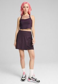 Fialový crop top s prekríženými ramienkami, kombinovaný s textúrovanou sukňou šortkami v rovnakom odtieni. Model má na sebe farebné športové topánky a biele ponožky.