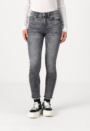 Person trägt ausgewaschene schwarze Skinny Jeans mit ausgefransten Säumen, weiße Plateausneaker mit Schnürung, schwarze Socken und ein transparentes weißes langärmeliges Oberteil.