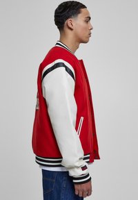 Rød varsity jakke med hvide læderærmer, sorte striber på skuldrene, ribbede manchetter og kontrasterende kant. Har trykknaplukning og sidelommer.