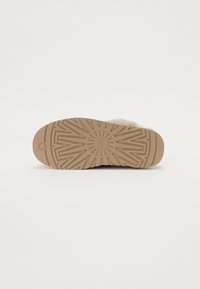 Semelle en caoutchouc d'une pantoufle avec des motifs de bande de roulement circulaires et en zigzag, couleur beige, et vue partielle de la partie supérieure fourrée.