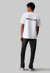 Camiseta de algodón blanca con cuello redondo, que presenta un gran logo negro de "Calvin Klein Jeans" en la parte posterior. Combinada con jeans gris oscuro.