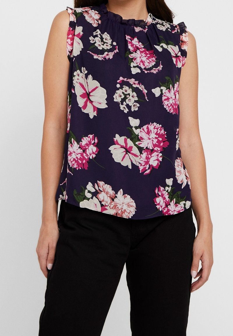 Blouse sans manches bleu marine à motifs floraux roses et blancs, avec un col à volants, un tissu léger et une coupe décontractée. Associée à un pantalon noir.