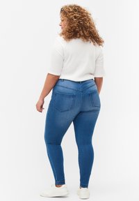 Jeans skinny de denim azul con cintura de altura media, bolsillos traseros y una textura lisa, combinados con una blusa blanca de manga corta y zapatillas blancas.